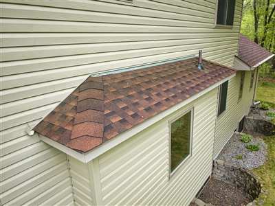 Close-up of a house with beige siding, featuring a small shingled roof over a window, surrounded by trees and greenery.